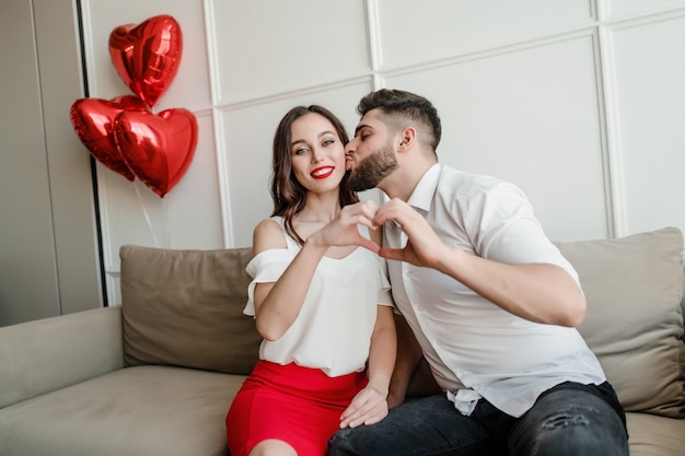 Handsome couple man and woman making heart shape with hands with red balloons sitting at home on couch