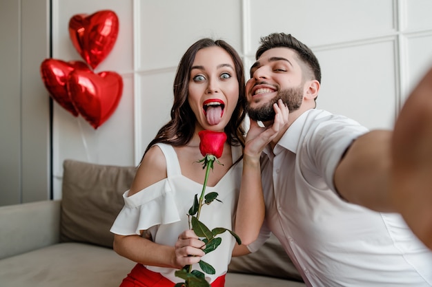 handsome couple man and woman making selfie with red rose and heart shaped balloons at home on couch