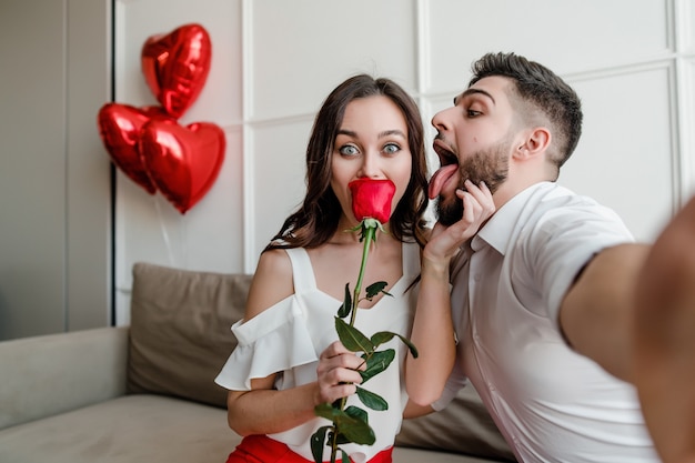 handsome couple man and woman making selfie with red rose and heart shaped balloons at home on couch