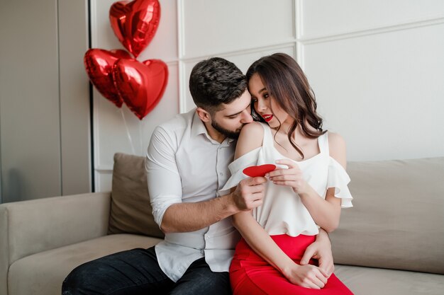 Photo handsome couple man and woman with valentine card and heart shaped balloons on couch at home