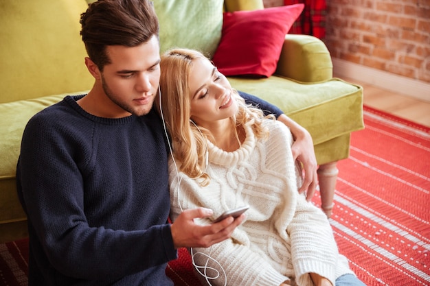 Handsome couple in sweaters sitting near the sofa with phone
