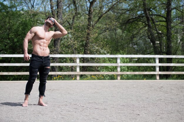 黒のジーンズとカウボーイハットを身に着けている屋外の牧場で強いポーズをとって立っているハンサムなカウボーイの男