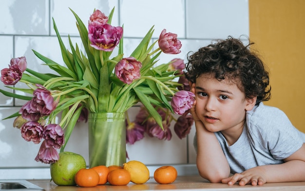 Handsome curly caucasian little boy leaning on kitchen table with vase of tulips and fruits