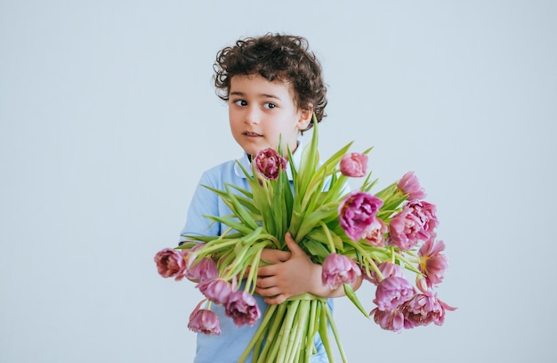 Handsome curly Italian little boy holds bouquet of stands against white wall looks aside