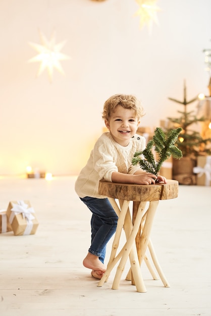 잘 생긴 곱슬 어린 소년은 화이트 룸에서 크리스마스 트리 근처 나무 의자에 기댈