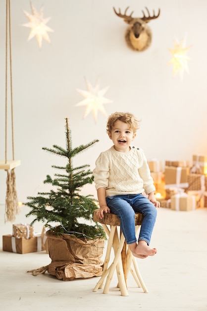 A handsome curly little boy sits on a wooden chair near a Christmas tree