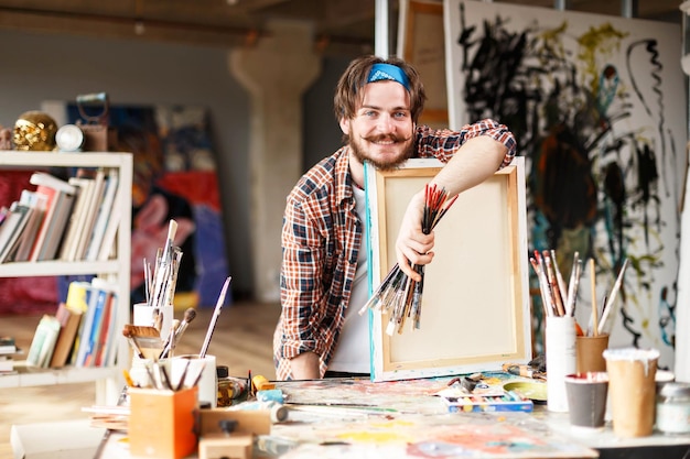 Handsome darkhaired bearded hipster male artist in blue bandana holding brush and leane on canvas in his contemporary studio with many paintings