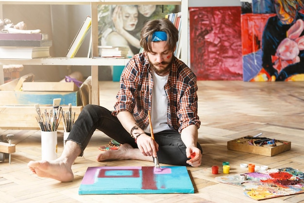 Handsome darkhaired bearded hipster male artist in blue bandana sitting on the floor and and looking on canvas in his contemporary studio with many paintings