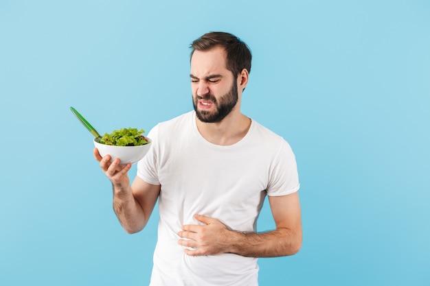 Handsome disgusted young bearded man wearing t-shirt standing isolated over blue wall, suffering from a stomach cramp