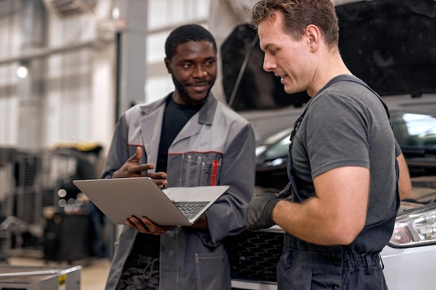 Handsome diverse mechanics in uniform examining car using laptop while working in auto service black...