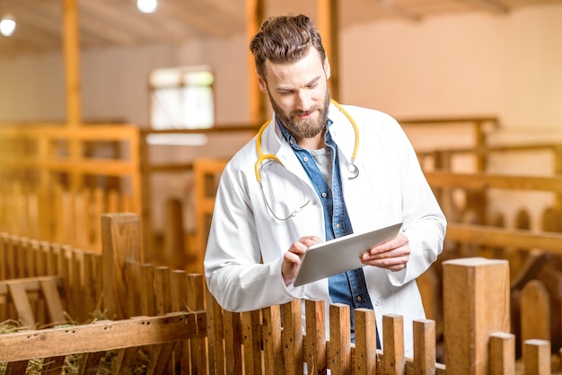 Handsome doctor in medical gown with digital tablet taking care about goats at the barn