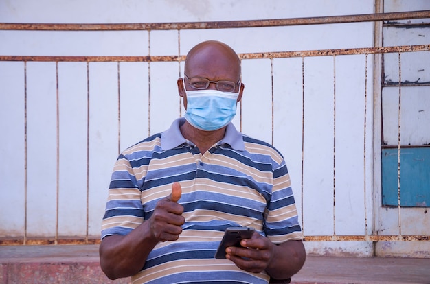 Un bell'uomo africano anziano che indossa una maschera per il viso che previene, si è impedito di scoppiare nella società sentendosi eccitato per ciò che ha visto sul suo cellulare e ha fatto il pollice in alto