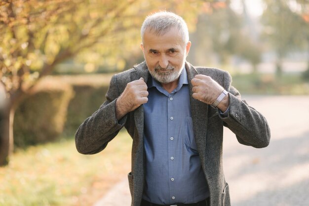 Handsome elderly man putting on a gray jacket Old grayhaired bearded man walk in the autumn park Yellow background