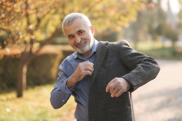 Handsome elderly man putting on a gray jacket Old grayhaired bearded man walk in the autumn park Yellow background