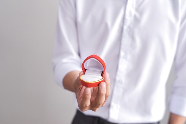 Handsome elegant man in a white shirt is holding a box with an engagement ring and shows in camera.