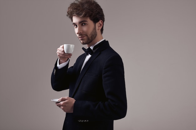 Uomo elegante bello con capelli ricci in smoking che tiene una tazza di caffè espresso