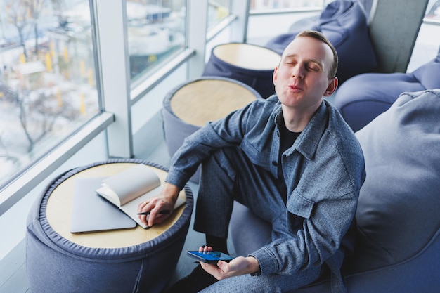 A handsome European young businessman in a coffee shop sits with a mobile phone and makes notes in a notebook Wearing an elegant casual suit and sitting with a laptop in a public cafe