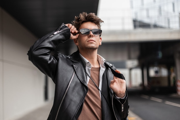 Handsome european young hipster man with trendy hairstyle in fashion leather black jacket straightens stylish sunglasses on a sunny spring day. Handsome guy in vintage clothes in retro style outdoors.