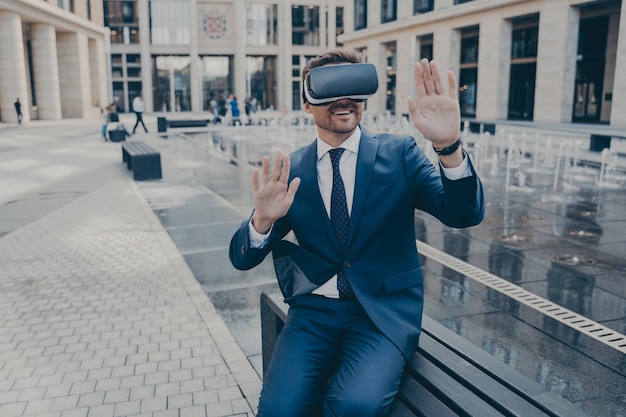 Handsome excited businessman sitting outside on bench while\
wearing portable virtual reality glasses, holding both of his hands\
in front of him, office buildings in blurred background
