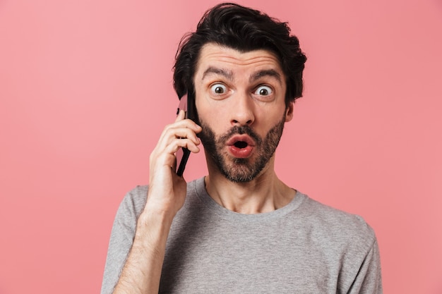 Handsome excited young bearded brunette man wearing sweater standing over pink, talking on mobile phone