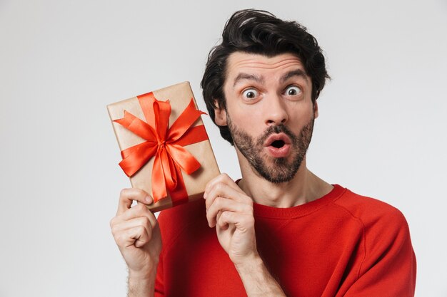 Photo handsome excited young bearded brunette man wearing sweater standing over white, holding present box