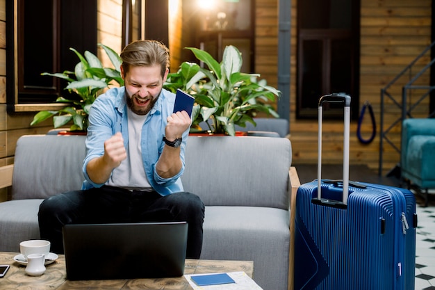 Handsome excited young man is sitting at sofa in hotel lobby with laptop and passport in hands