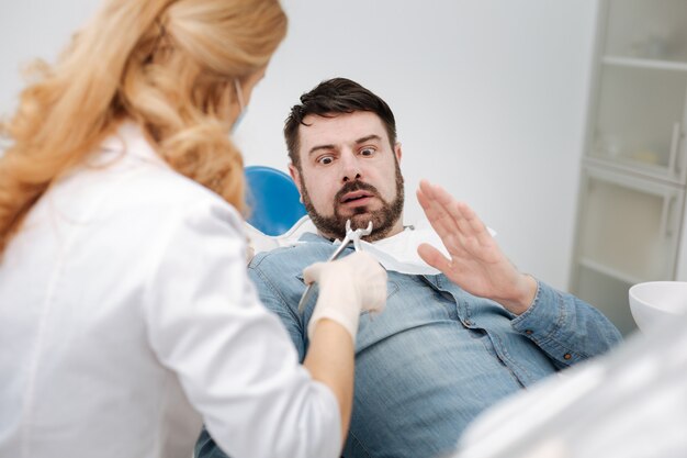 Handsome expressive young man looking scared and unwilling having his teeth out while paying a visit to his doctor