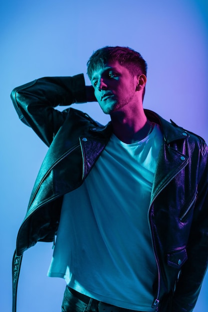 Handsome fashionable brutal man with a hairstyle in fashion outerwear with a leather jacket poses in the studio with blue and pink light. Colorful Art portrait of a guy