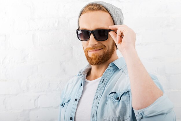 Handsome and fashionable. Confident young bearded man adjusting his eyewear and looking at camera while standing against brick wall