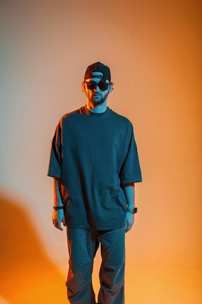 Handsome fashionable guy with a cap in a trendy fashion tshirt and pants in a studio with orange and neon light