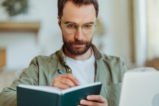 Handsome focused bearded businessman wearing eyeglasses taking notes studying at home