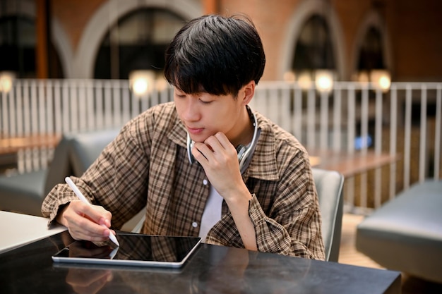 Handsome and focused young Asian man taking notes on a digital tablet remote working at cafe