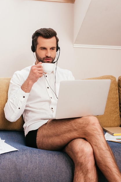 Handsome freelancer in shirt and panties drinking coffee and using headset with laptop on couch