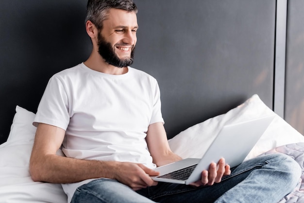 Handsome freelancer smiling while holding laptop on bed