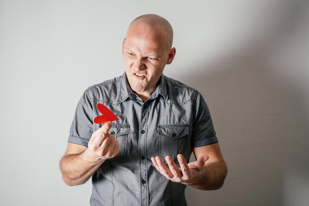 Handsome frowning bald middle-aged man in gray shirt hold red paper heart with two fingers indignantly with hatred on white background. Valentine's day, love, hatred emotion concept. Copy space.