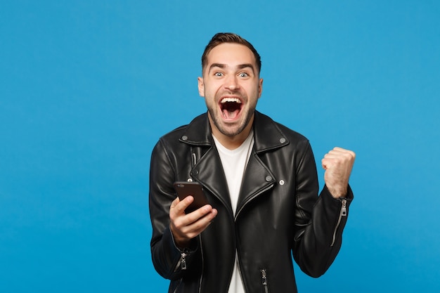 Handsome fun overjoyed excited young unshaven man in black leather jacket white t-shirt using mobile phone isolated on blue wall background studio portrait. People lifestyle concept Mock up copy space