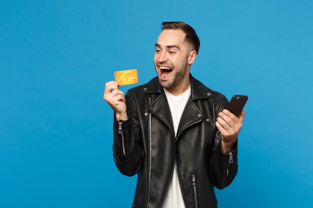 Bello divertente elegante giovane uomo con la barba lunga in giacca nera t-shirt bianca tenere in mano il cellulare, carta di credito isolata sul ritratto in studio di sfondo muro blu. concetto di stile di vita della gente. mock up copia spazio