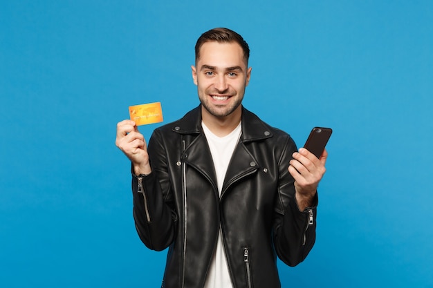 Handsome fun stylish young unshaven man in black jacket white t-shirt hold in hand cellphone, credit card isolated on blue wall background studio portrait. People lifestyle concept. Mock up copy space