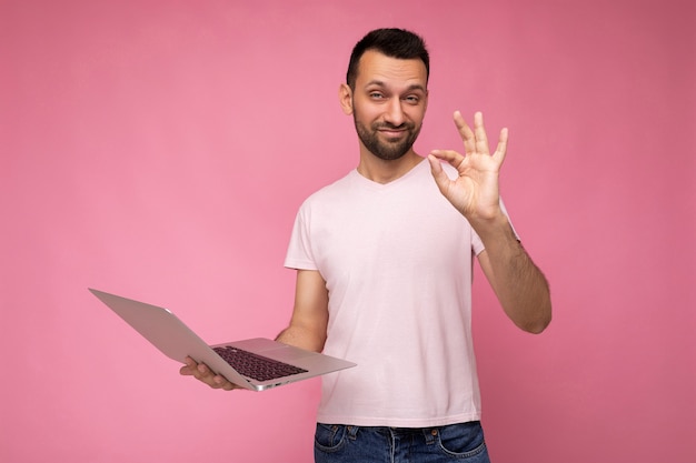 Uomo divertente bello del brunet che tiene il computer portatile e che mostra il gesto va bene guardando la fotocamera in t-shirt sulla superficie rosa isolata