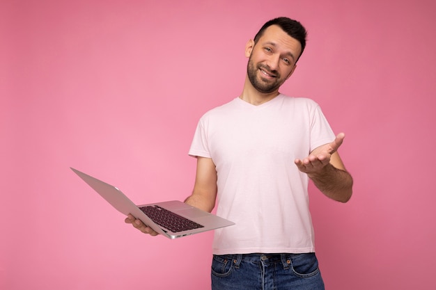 Handsome funny and smiling brunet man holding laptop computer and showing hand  in t-shirt on isolated pink wall.