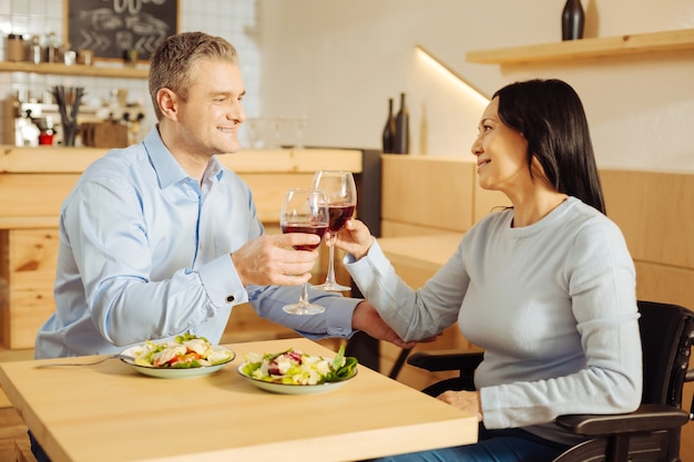 Handsome gleeful well-built man and a pretty inspired handicapped woman smiling and drinking wine and having dinner