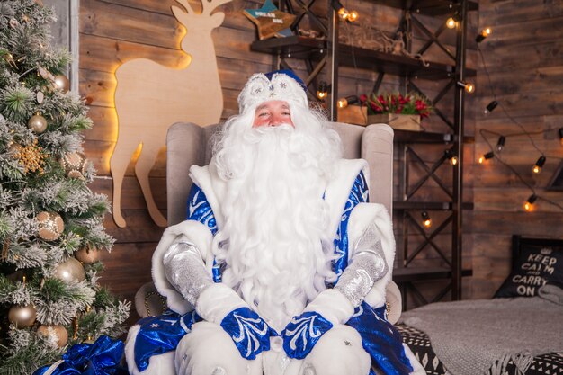 Handsome Grandfather Frost in a blue fur coat sits near a Christmas tree