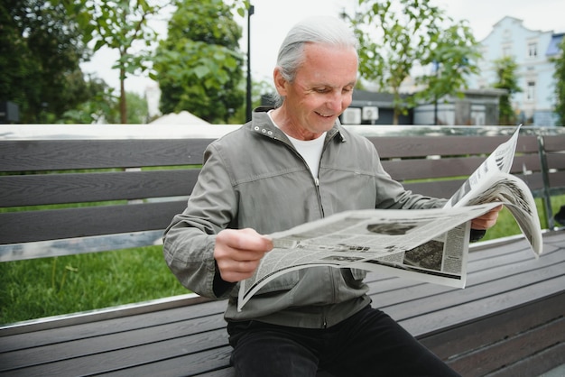 Красивый дедушка сидит на скамейке в парке и читает газету Старший седой мужчина