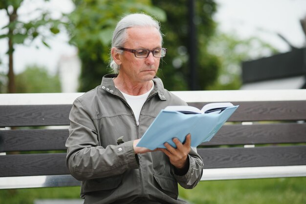 Handsome grandfather sits on a bench in the park and reads a newspaper Senior grayhaired man