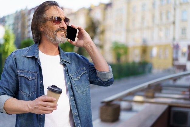 Handsome grey beard middle aged man talking on the phone holding coffee in disposable paper cup standing outdoors in old city background wearing jeans shirt Freelancer traveling man