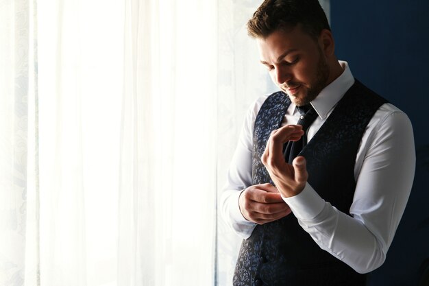 Handsome groom in stylish blue waistcoat stands before a window and fixes his cufflinks