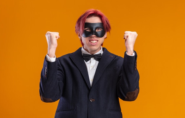 Handsome groom in suit wearing bow tie and masquerade mask\
happy and excited clenching fists standing over orange wall