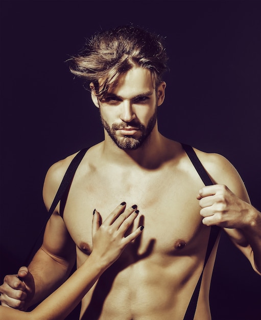 Handsome guy fashion sexy young bearded macho man model with suspenders on pants with female hand on bare muscular torso holds on grey surface