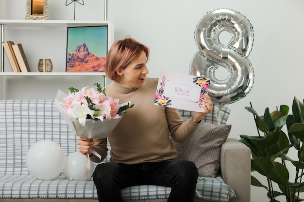 Handsome guy on happy women day holding bouquet looking at postcard in his hand sitting on sofa in living room