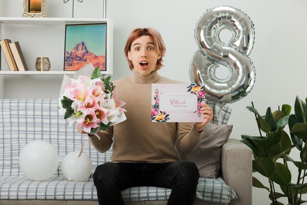 Handsome guy on happy women day holding bouquet with postcard sitting on sofa in living room